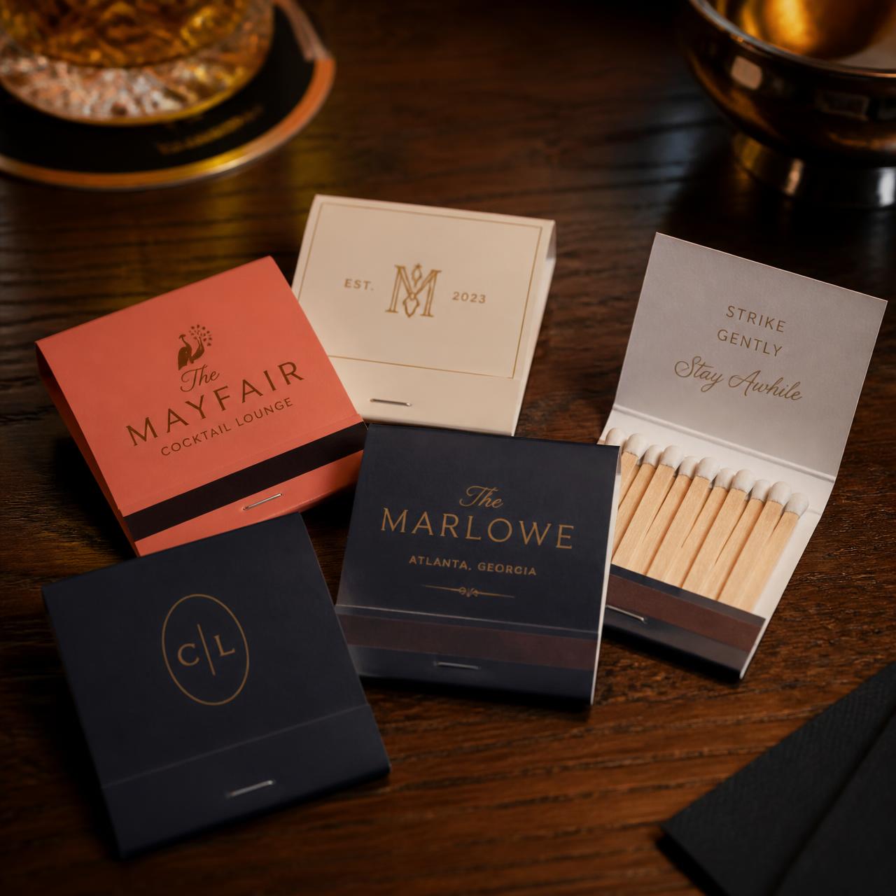 Custom printed matchbooks in coral, cream and navy with gold logos on a walnut bar counter, one open showing wood-stem matches